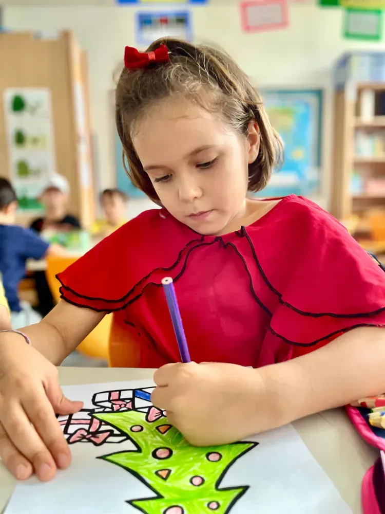 A little girl is sitting at a table in a classroom drawing a christmas tree.