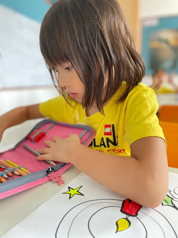 A little girl in a yellow shirt is sitting at a table with a pencil case filled with colored pencils.