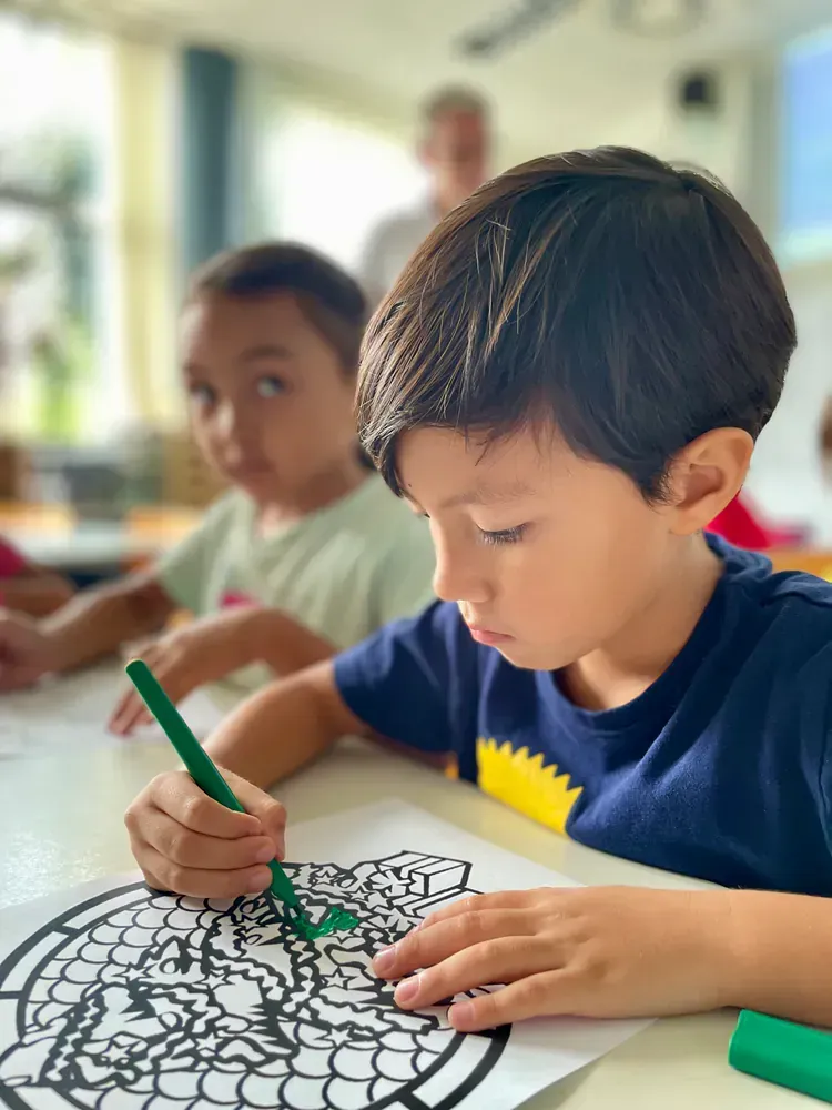 A young boy is sitting at a table drawing with a green marker.
