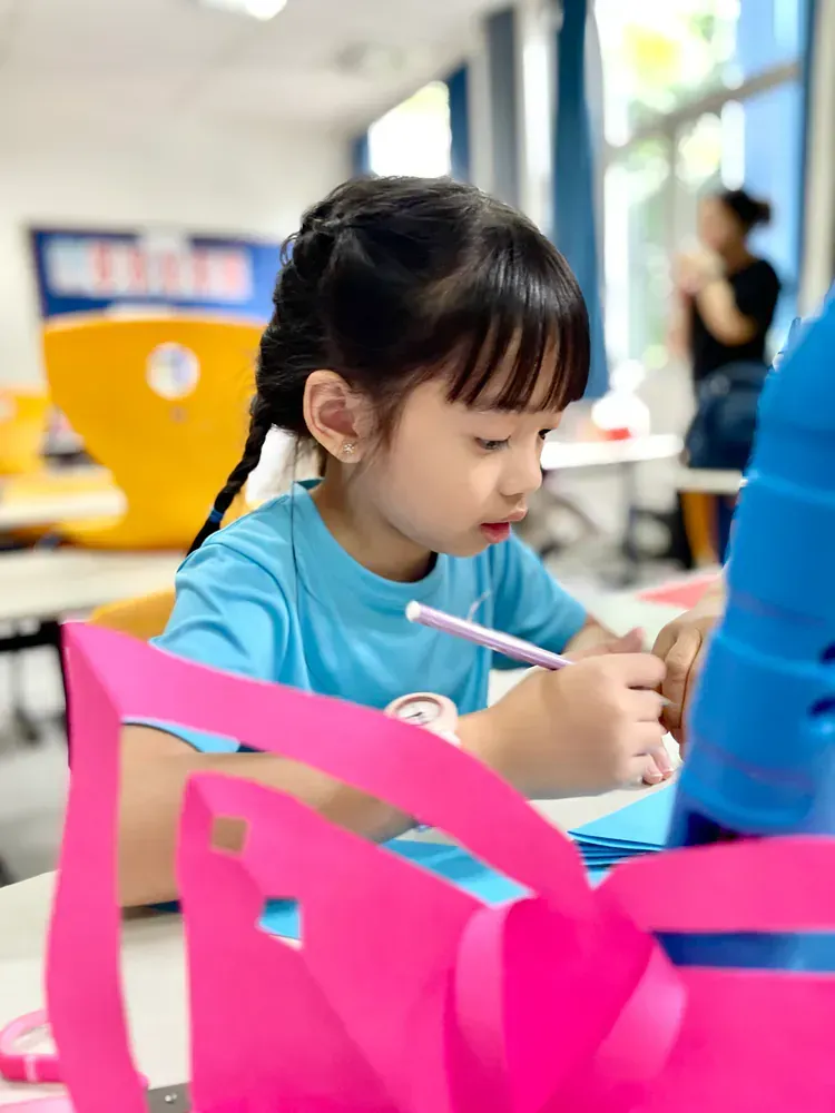 A little girl is sitting at a table with a pink ribbon and a pen.