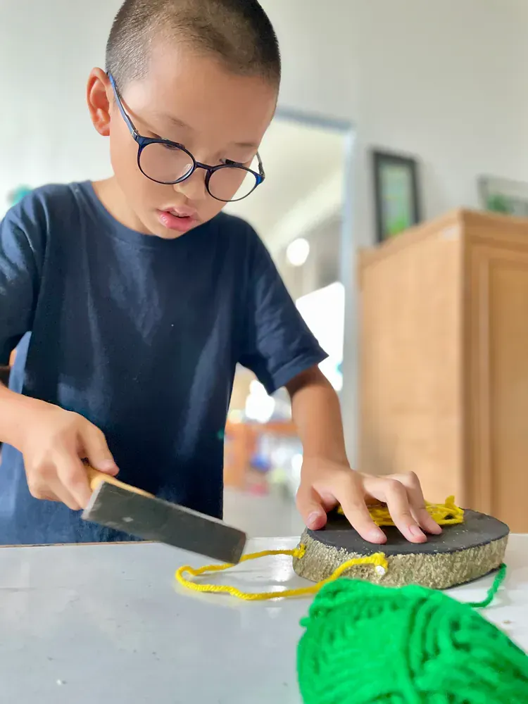 Boy wearing glasses and a navy blue shirt carefully hammering a small nail into an object
