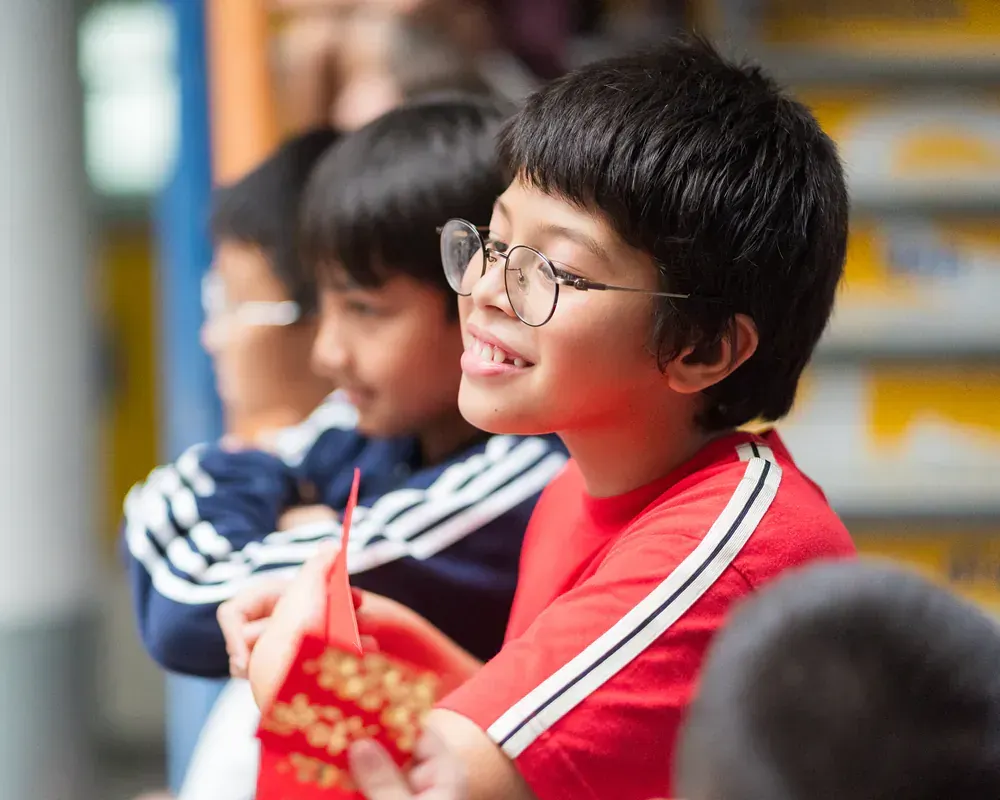A boy wearing glasses and a red shirt is holding a red envelope.