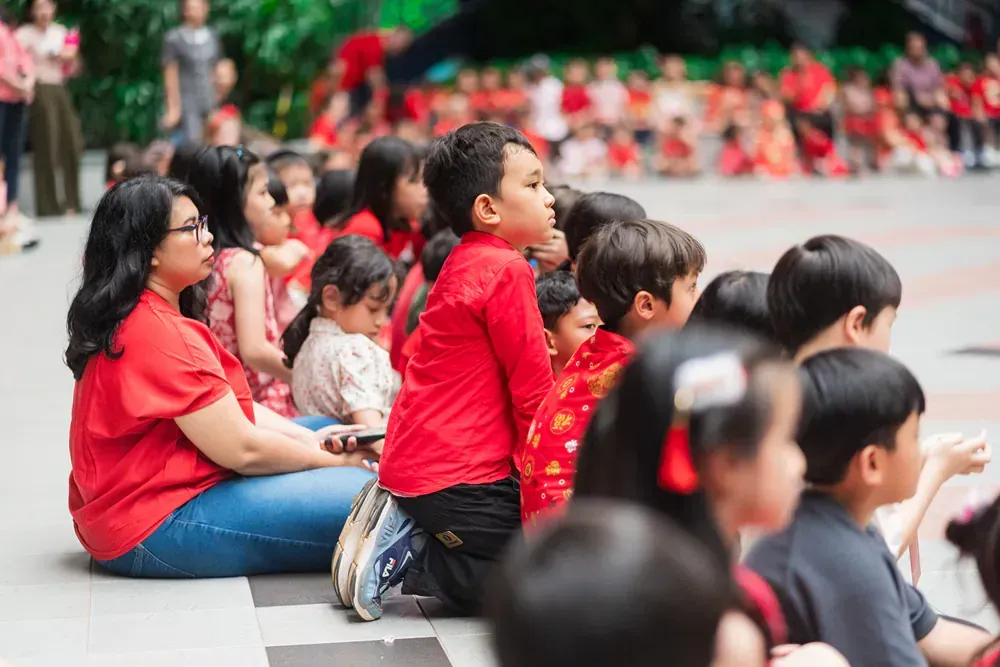 A group of children are sitting on the ground with a woman in a red shirt.