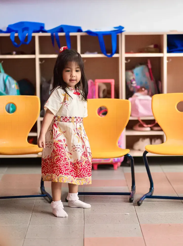 A little girl in a dress is standing in front of yellow chairs in a classroom.