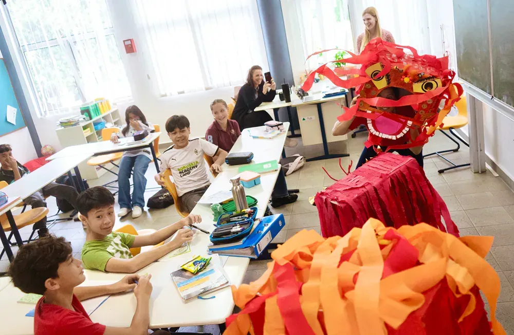 A group of children are sitting at tables in a classroom with a dragon costume on.