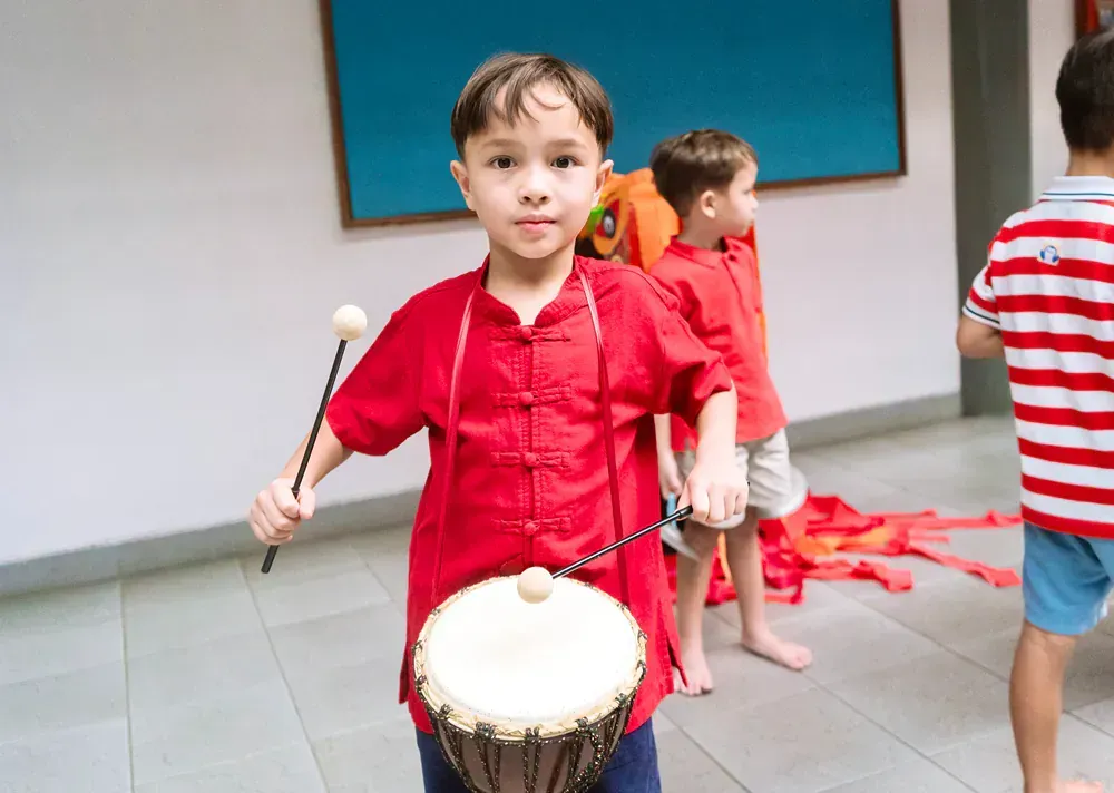 A young boy in a red shirt is playing a drum with a stick.