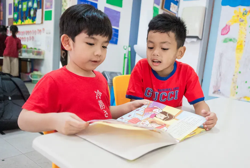 Two young boys are sitting at a table reading a book.