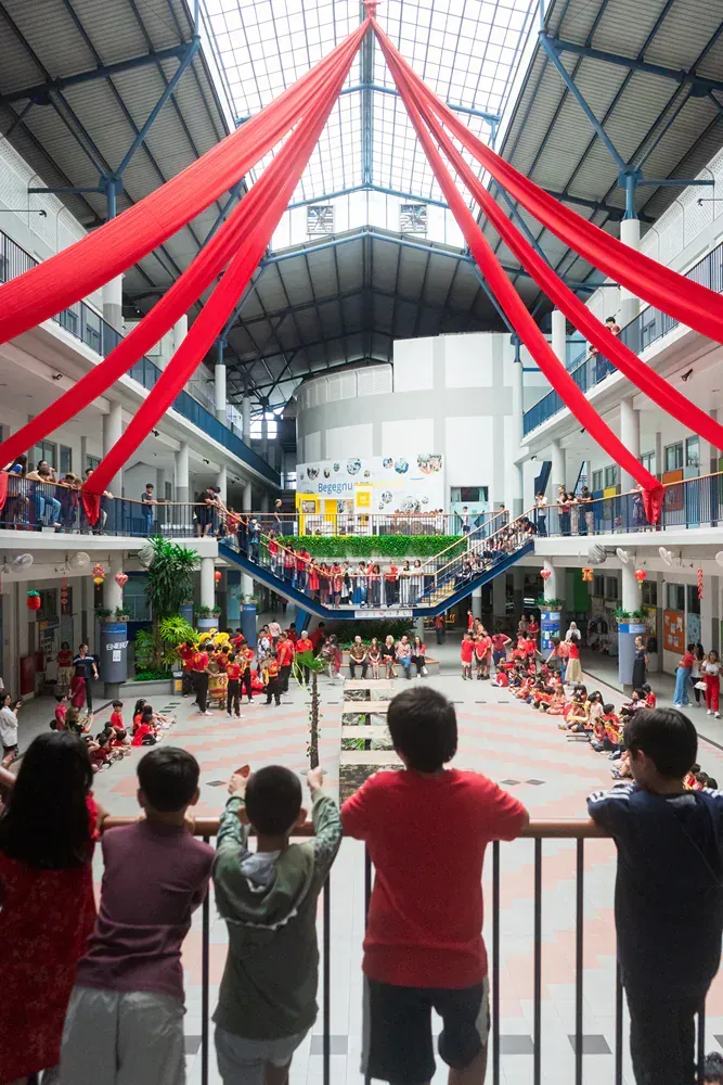 A group of children are standing on a balcony looking at a large building with red ribbons hanging from the ceiling.