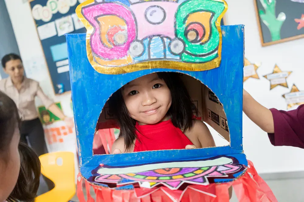 A little girl is looking out of a cardboard box in a classroom.