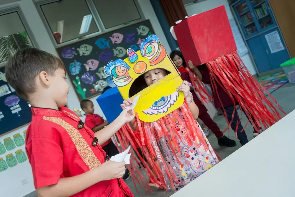 A boy in a red shirt is holding a lion mask in a classroom.