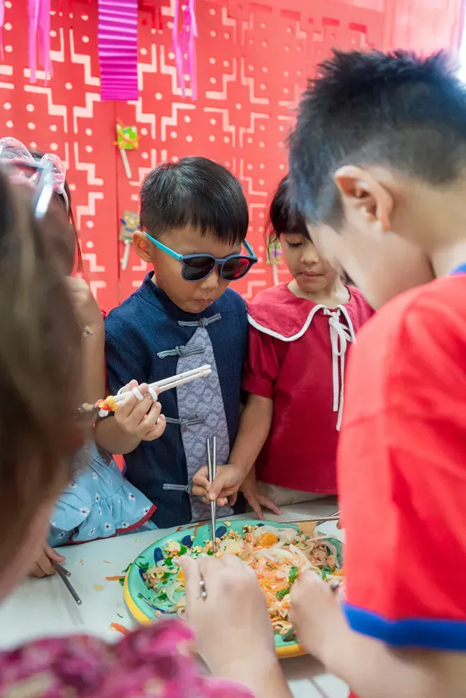 A group of children are sitting around a table eating food with chopsticks.