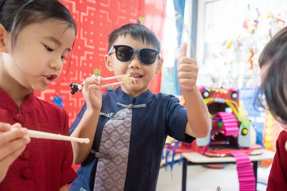 A boy wearing sunglasses is eating food with chopsticks and giving a thumbs up.