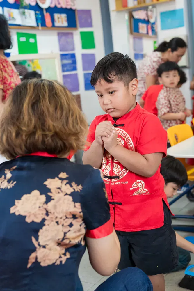 A woman is kneeling down next to a boy in a red shirt in a classroom.