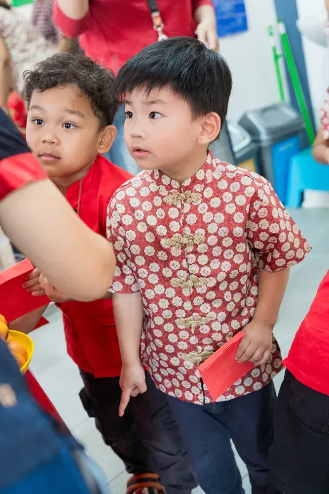 Two young boys are standing next to each other in a room holding red envelopes.