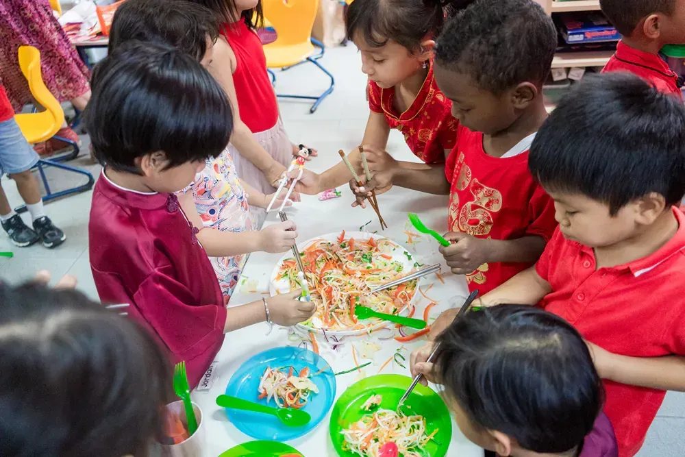 A group of children are sitting around a table eating food with chopsticks.
