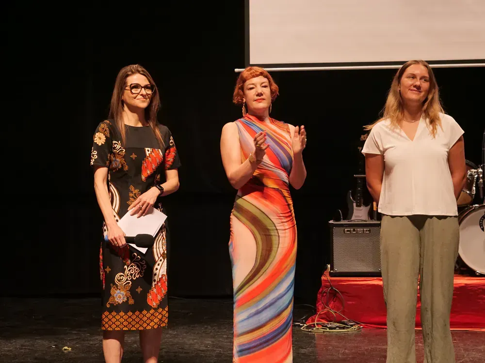 Three women standing on a stage with one wearing a colorful dress