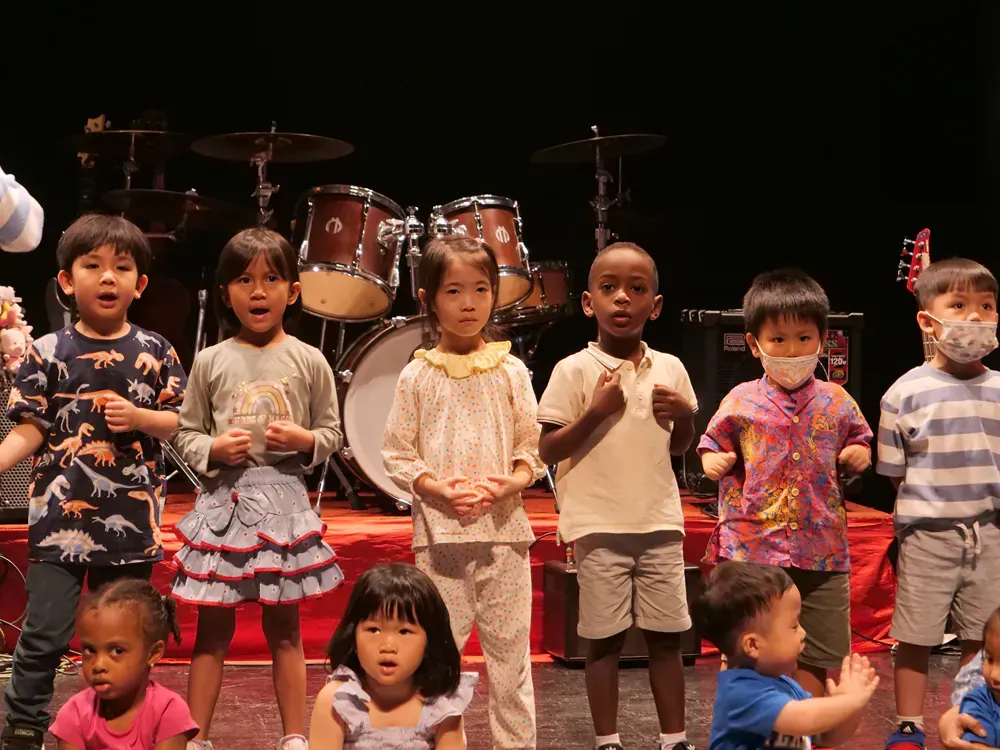A group of children are standing on a stage in front of a drum set.