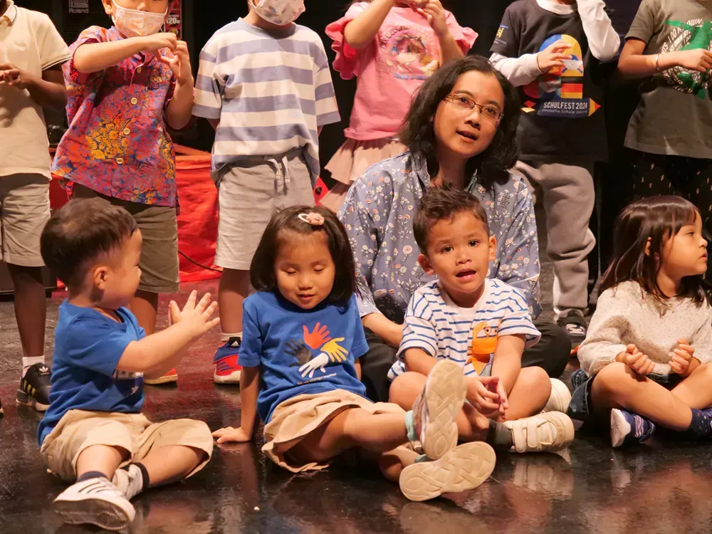 A group of children are sitting on the floor with a woman standing behind them.
