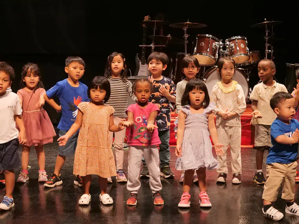 A group of children are standing on a stage in front of a drum set.