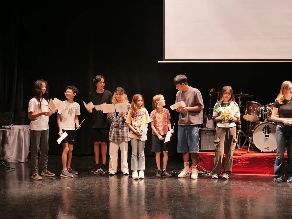 A group of children are standing on a stage reading papers