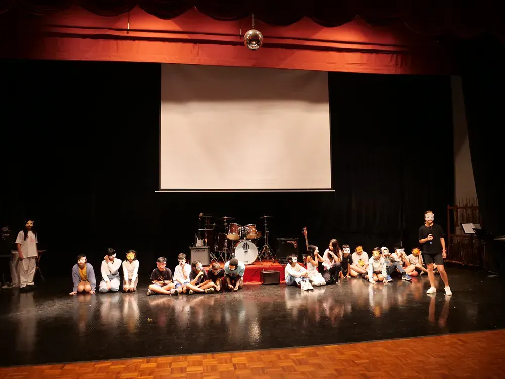 A group of people are sitting on the floor on a stage in front of a projector screen.