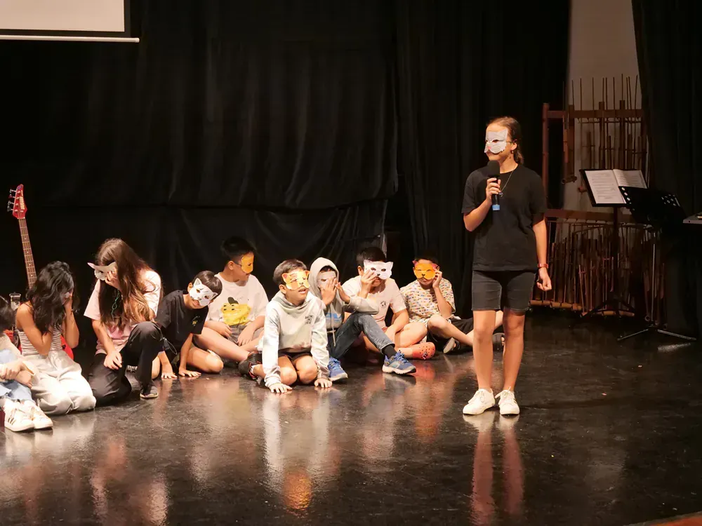 A group of children wearing masks are sitting on the floor on a stage.