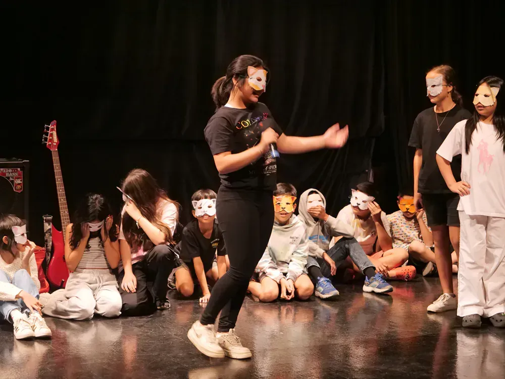 A group of children wearing masks are sitting on the floor on a stage.