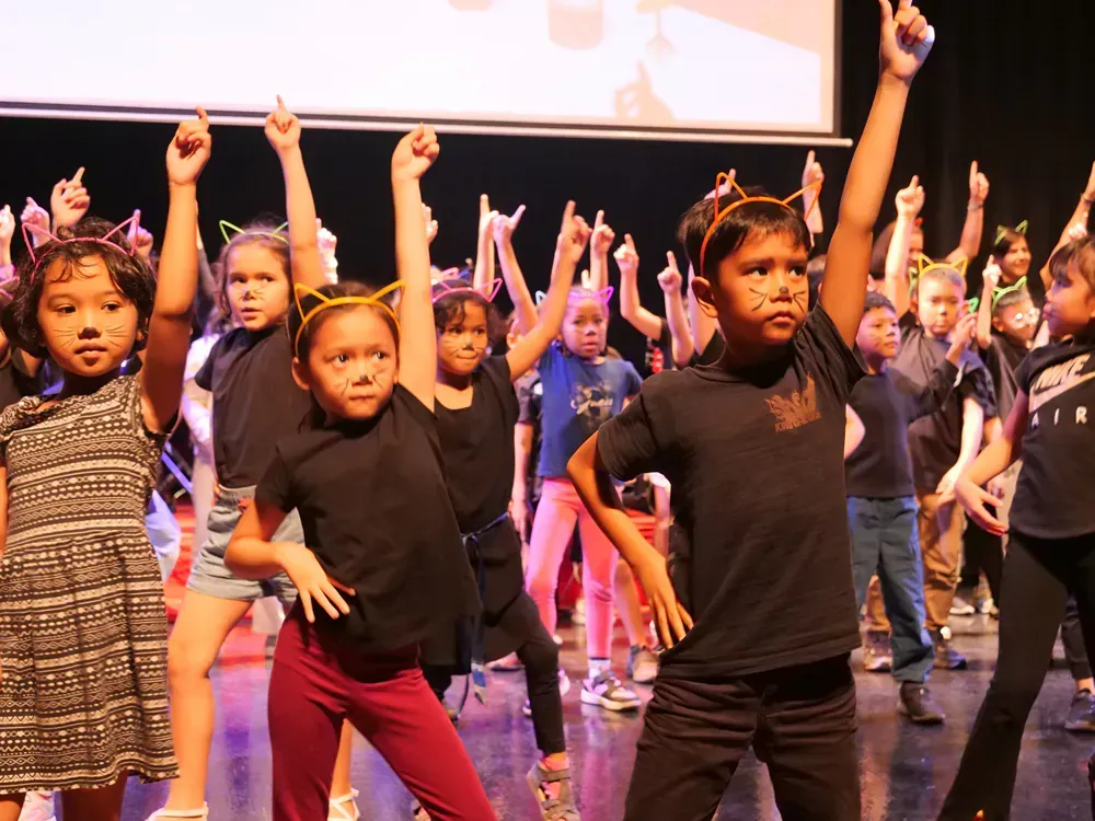 A group of children are dancing on a stage with their arms in the air