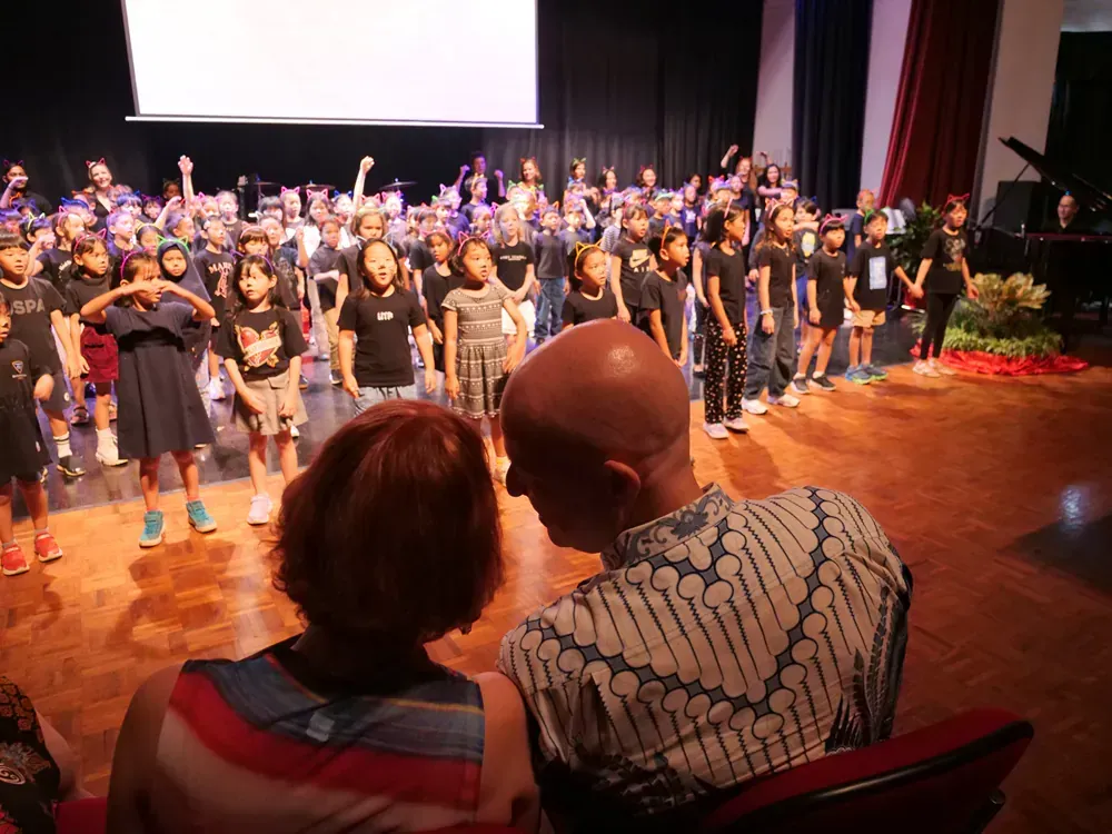 A man and woman are sitting in front of a large group of children on a stage.
