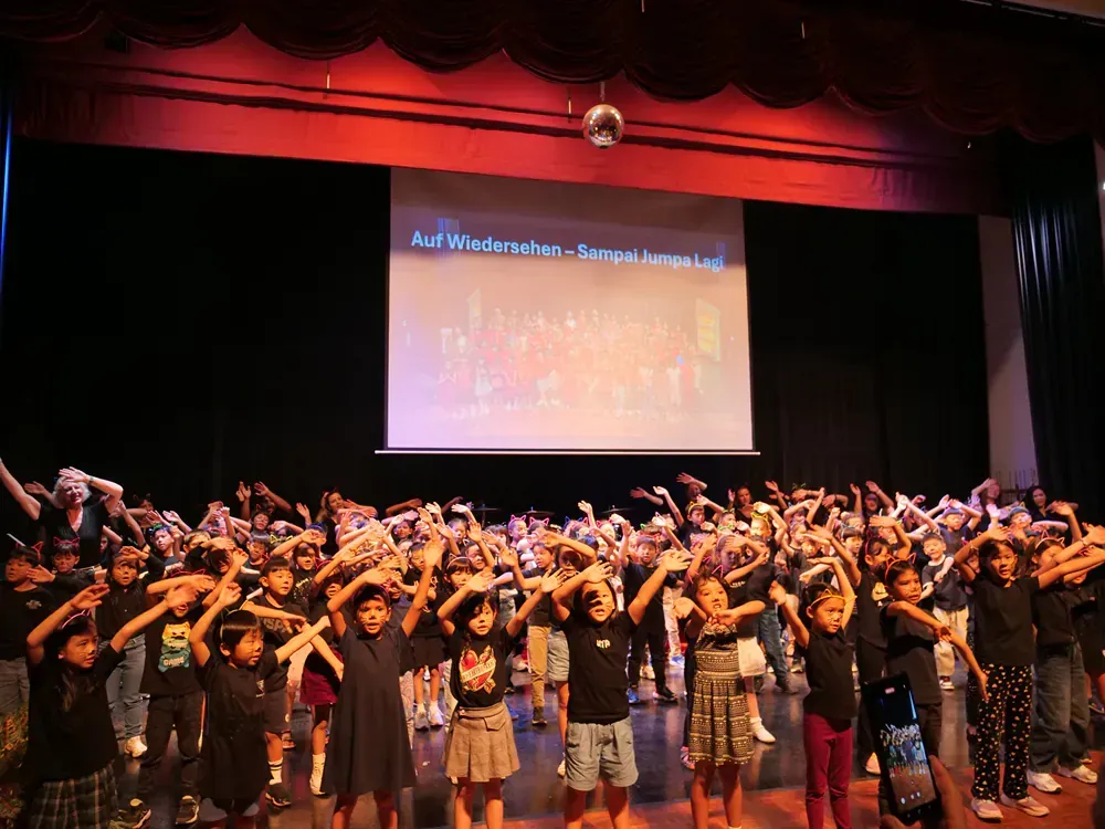 A large group of children are dancing on a stage in front of a large screen.