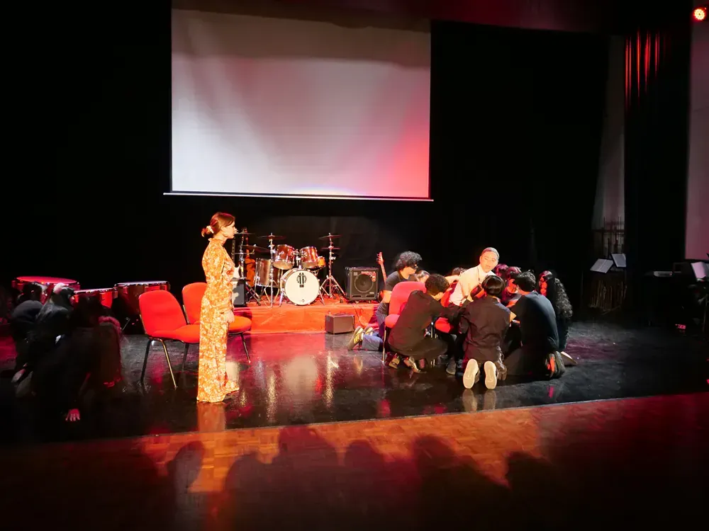 A group of people are sitting on the floor on a stage in front of a projector screen.