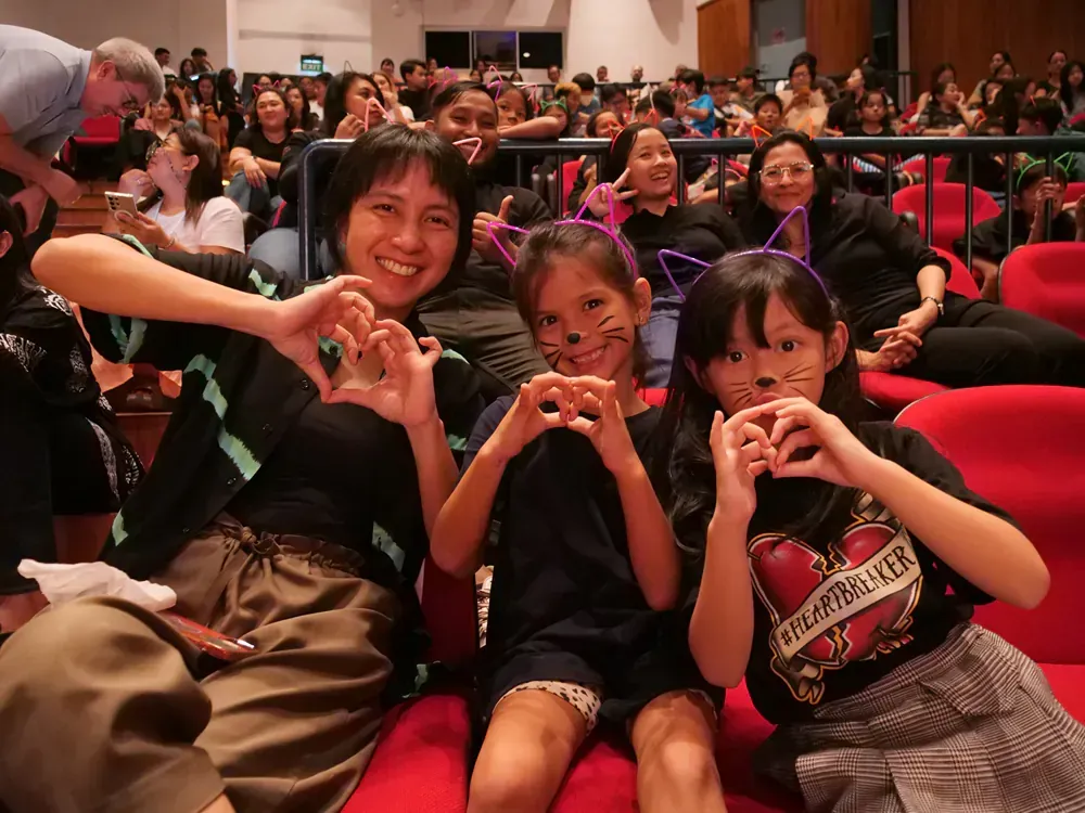 A group of people are sitting in a theater making hearts with their hands.
