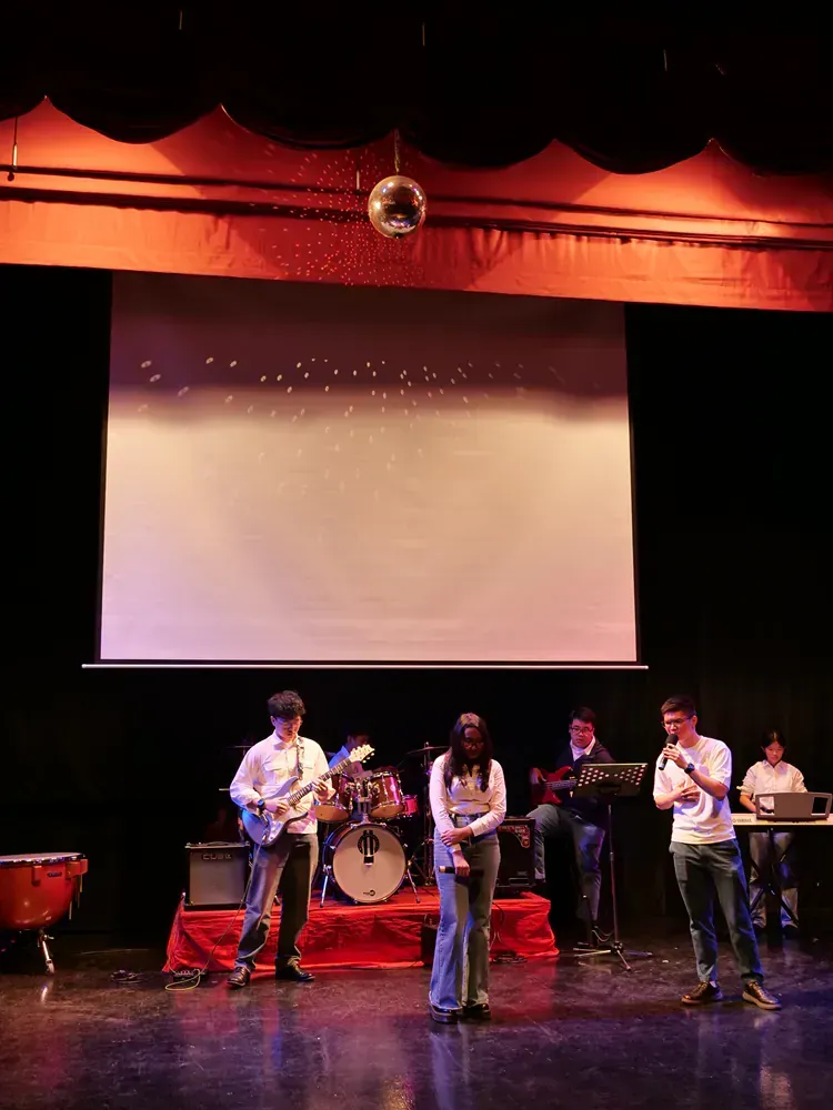 A group of people are playing instruments on a stage in front of a projector screen.