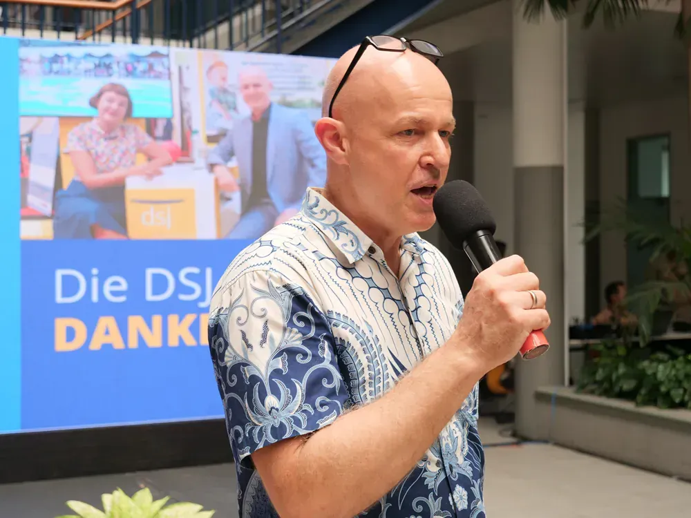 A bald man is speaking into a microphone in front of a sign that says die dsj danke