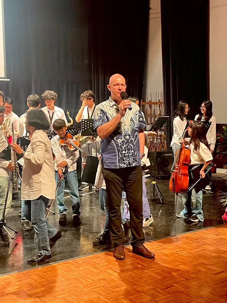 A man is standing on a stage holding a microphone in front of a group of children playing instruments.