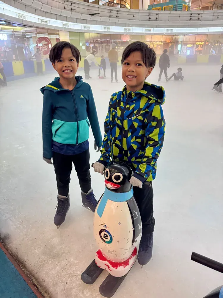 Two young boys are standing next to a penguin statue on an ice rink.