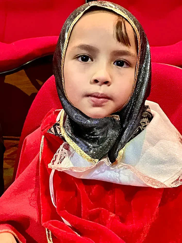 A little girl wearing a scarf and a red dress is sitting in a red chair.