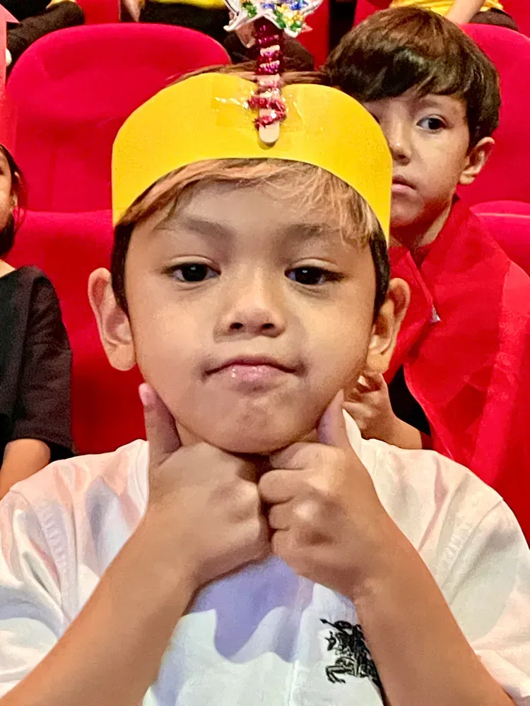 A young boy wearing a yellow crown is giving a thumbs up.