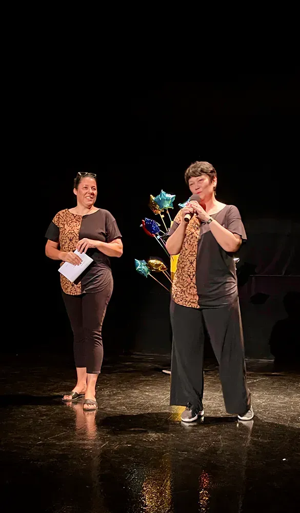 Two women are standing on a stage holding balloons and a microphone.