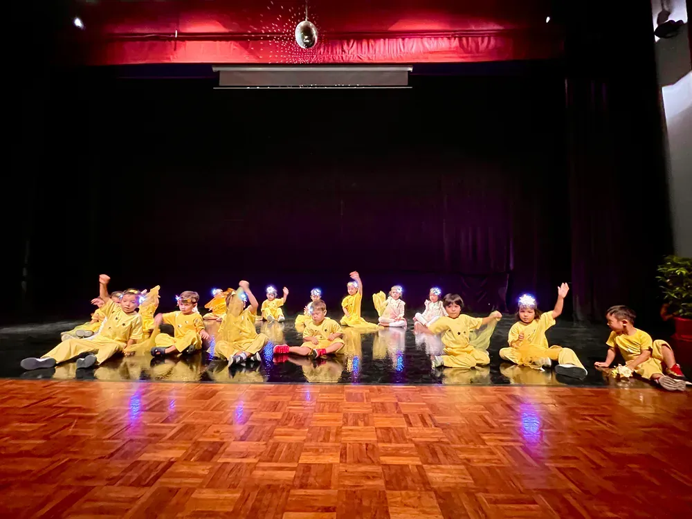 A group of children are sitting on a wooden dance floor on a stage.