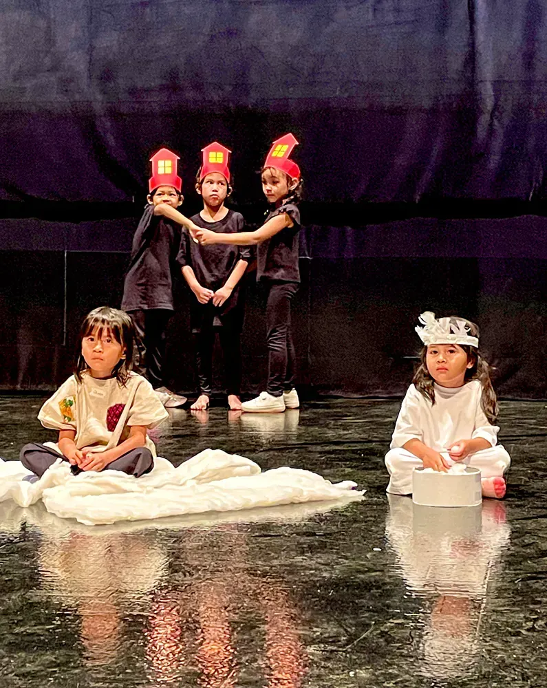 A group of children are sitting on a stage in front of a purple curtain.