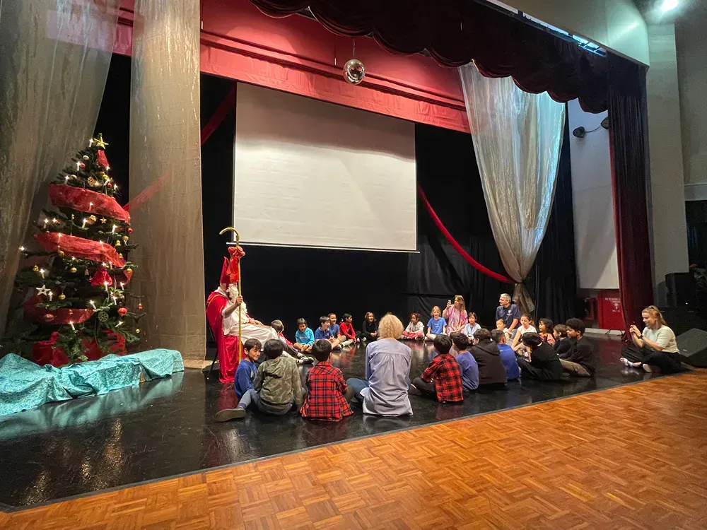 A group of children are sitting on the floor in front of a stage with a christmas tree.