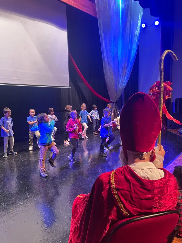 A group of children are dancing on a stage with a man in a red robe holding a cane.