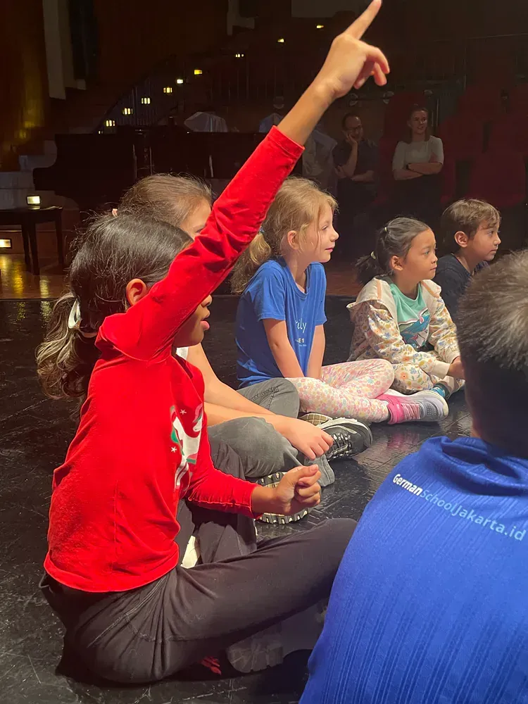A group of children are sitting on the floor and one of them is raising her hand.