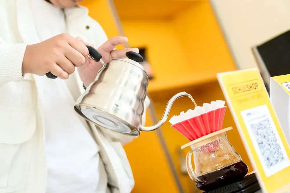 A person is pouring coffee from a kettle into a cup.