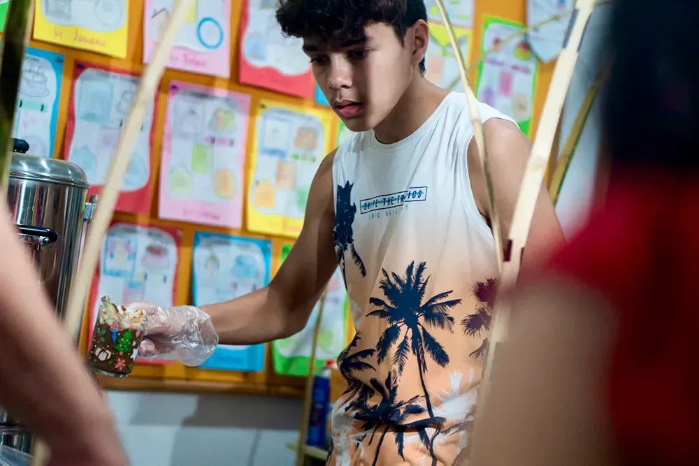 A young man wearing a tank top with palm trees on it is standing in front of a pot.