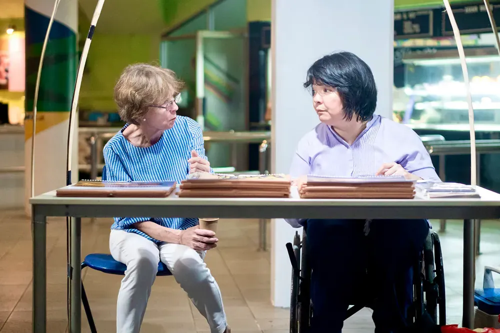 A woman in a wheelchair is sitting at a table talking to another woman.