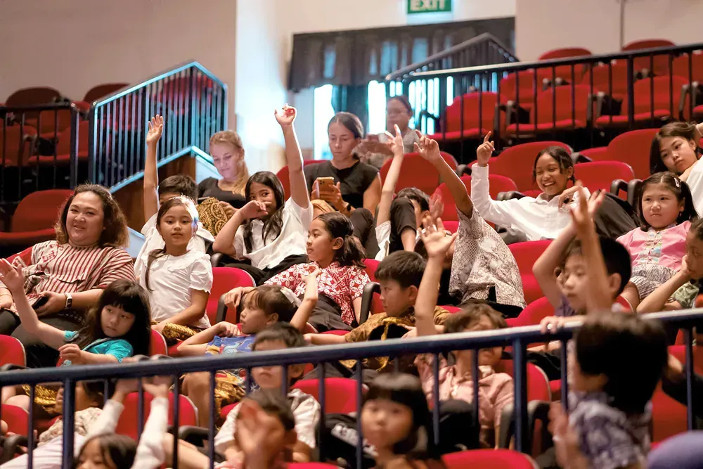 A group of children are raising their hands in an auditorium.