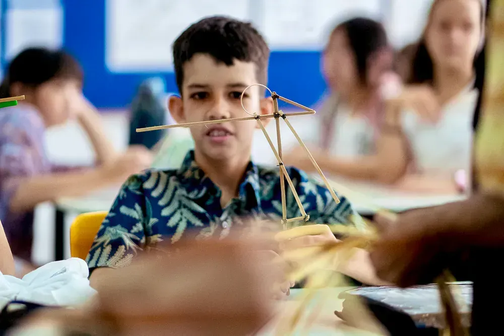 A young boy is sitting at a desk in a classroom making a structure out of sticks.
