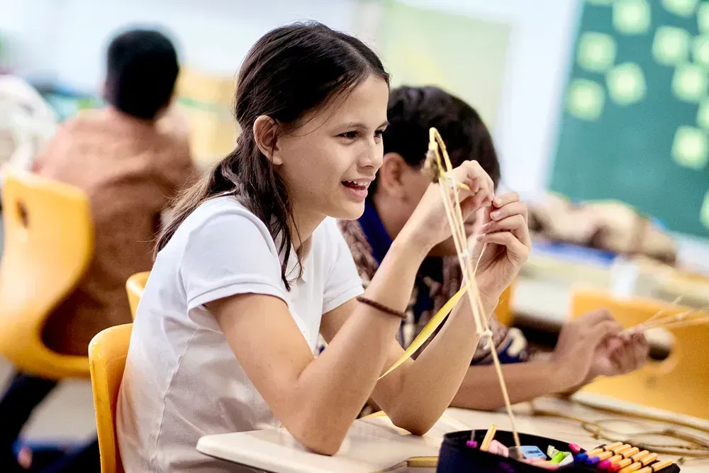 A girl is sitting at a desk in a classroom holding a piece of string.